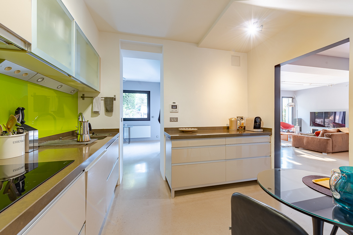 Modern kitchen with white cabinets, lime green backsplash, and stainless countertops; dining table and living room visible through an open doorway.