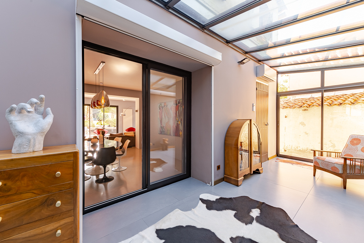 Sunlit indoor–outdoor living space with glass sliding doors, cowhide rug, and mid-century furniture pieces including a wooden dresser and vintage cabinet.