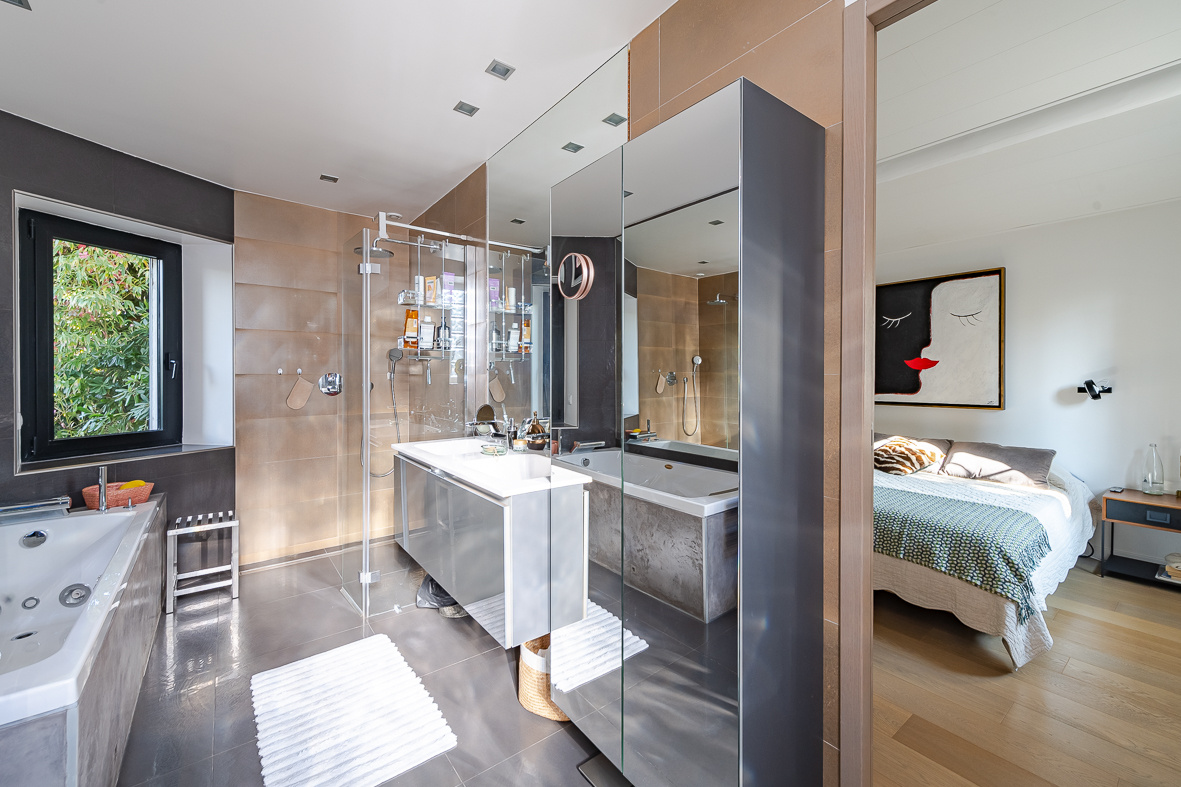 Luxurious bathroom with glass shower enclosure, freestanding tub, and a wide mirror wall; bedroom visible through the doorway.