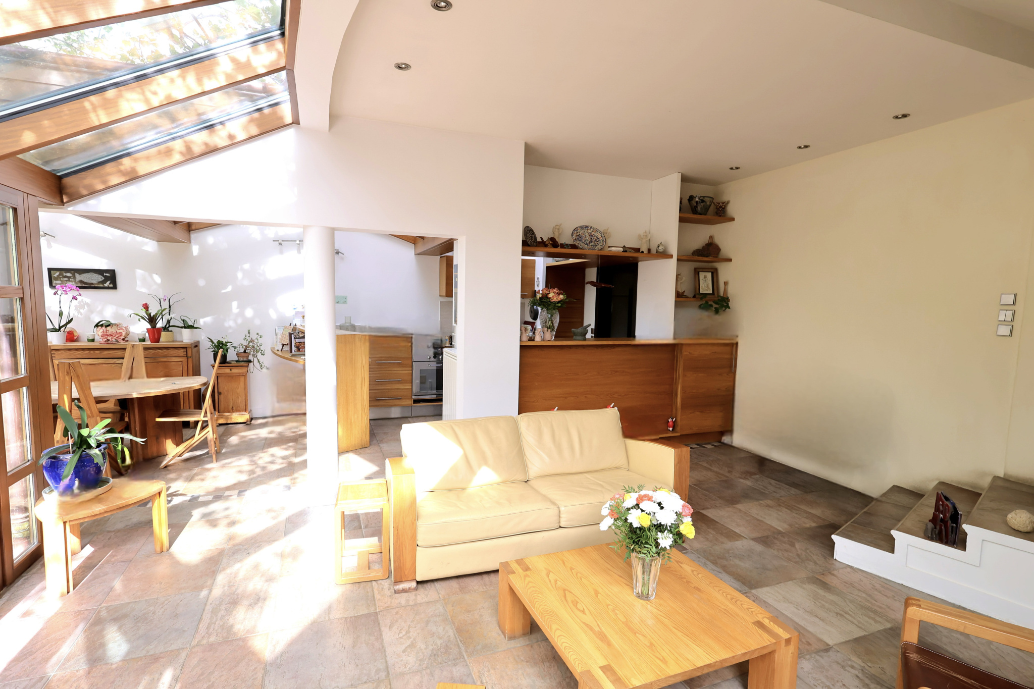 Open-plan living area with a cream sofa, wooden coffee table, and a vase of flowers; kitchen and dining area in the background to the left and back.