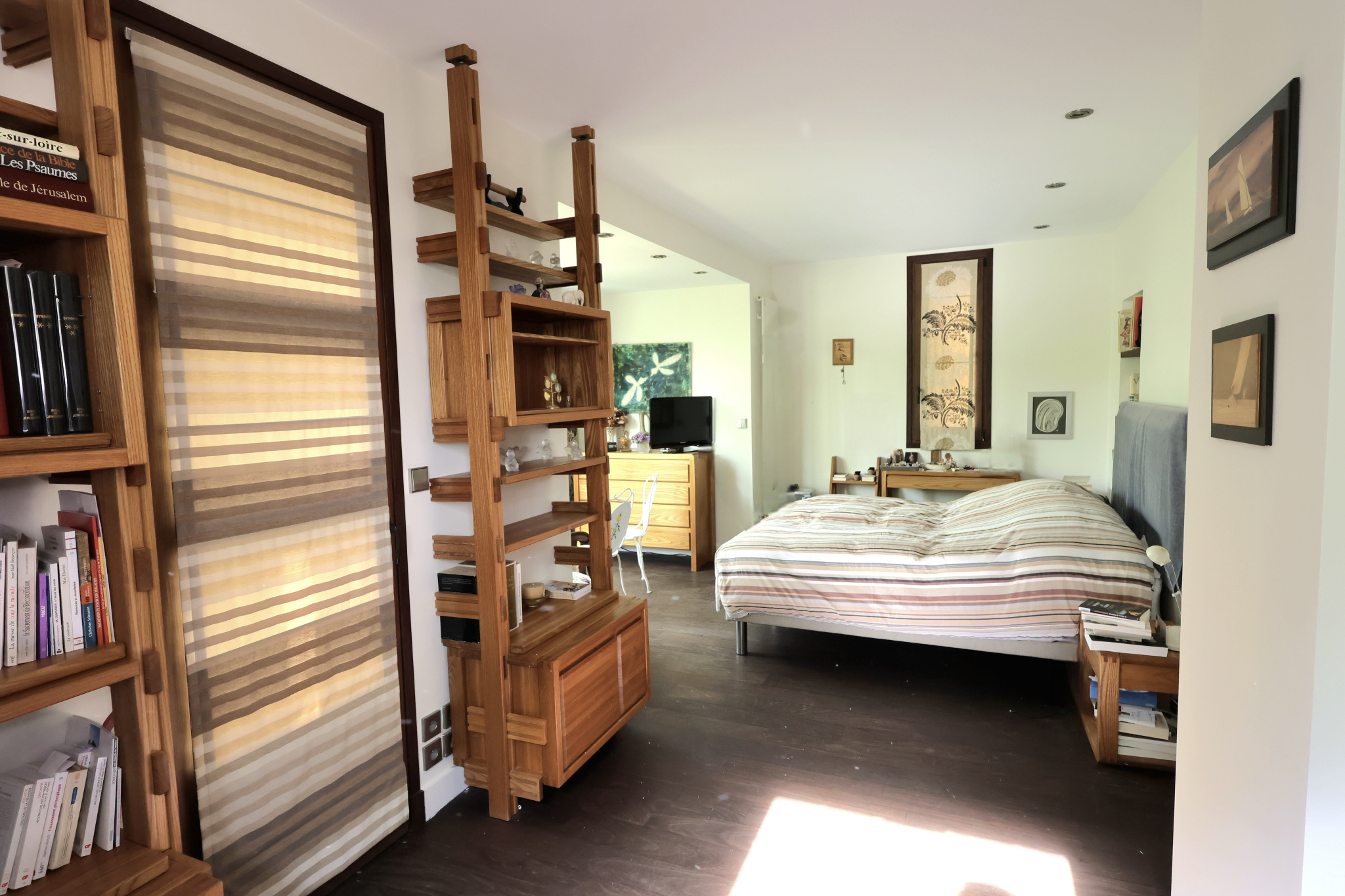 Bright bedroom with wooden modular shelves acting as a room divider, a striped bed with headboard, and a dresser with a small TV in the background.