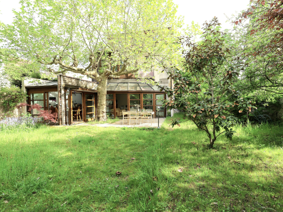 Backyard garden with a glass-walled patio and wooden outdoor dining set under a large leafy tree.