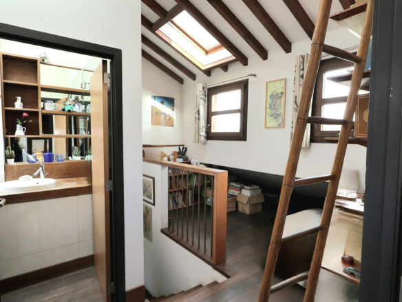 Cozy attic loft with exposed wooden beams, a tall wooden ladder, and a railing overlooking a study and bed area; skylight brings in light.