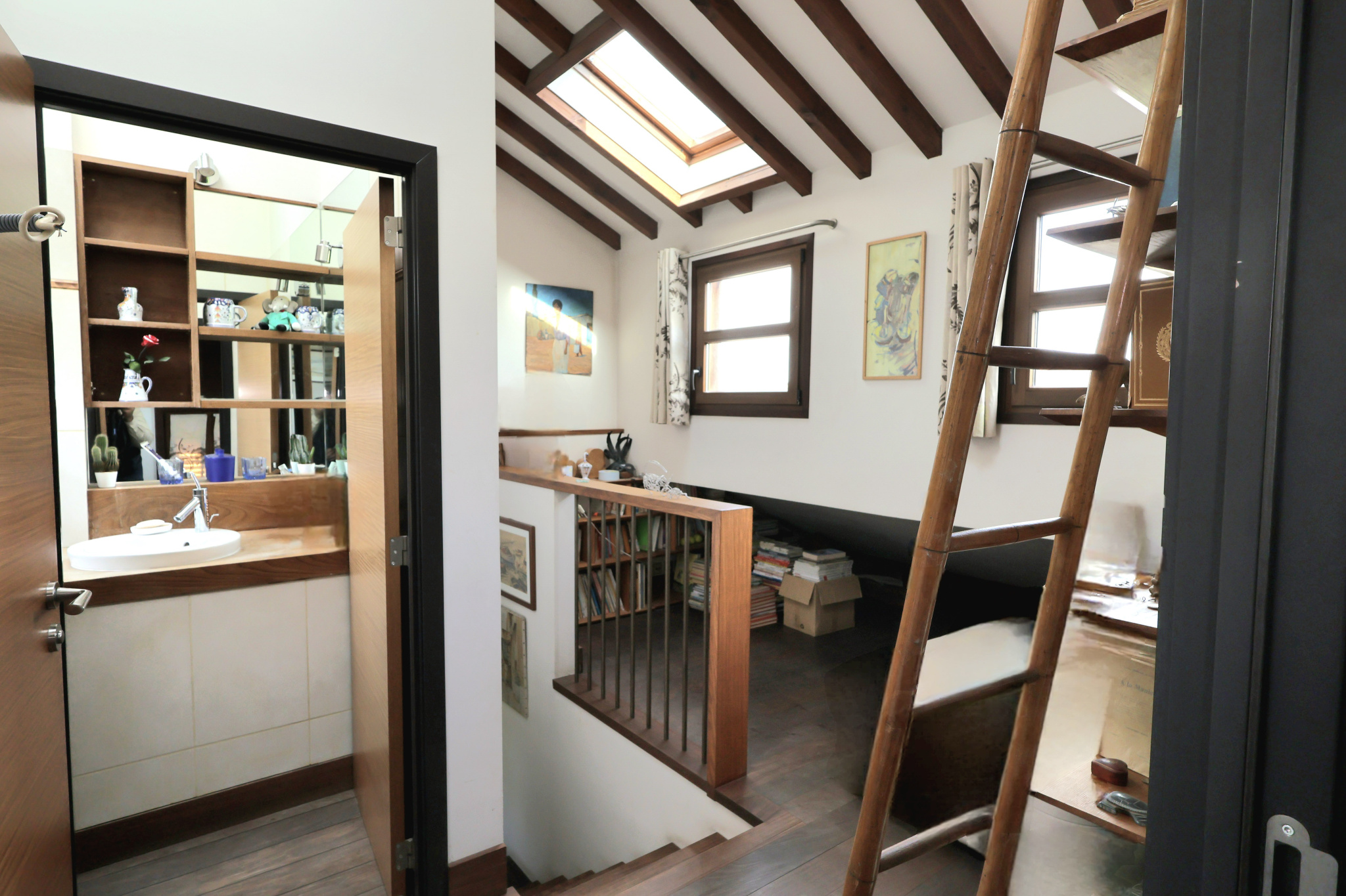 Cozy attic loft with exposed wooden beams, a tall wooden ladder, and a railing overlooking a study and bed area; skylight brings in light.