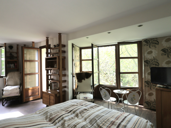 Sunlit bedroom with large windows revealing green trees, and a wooden shelving unit flanking a chair and bed in the foreground.