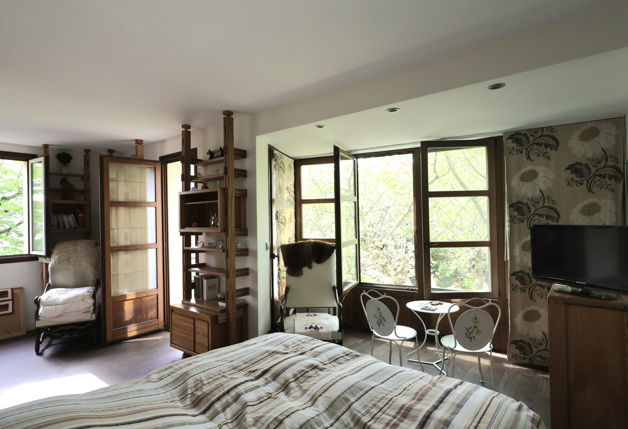 Sunlit bedroom with large windows revealing green trees, and a wooden shelving unit flanking a chair and bed in the foreground.