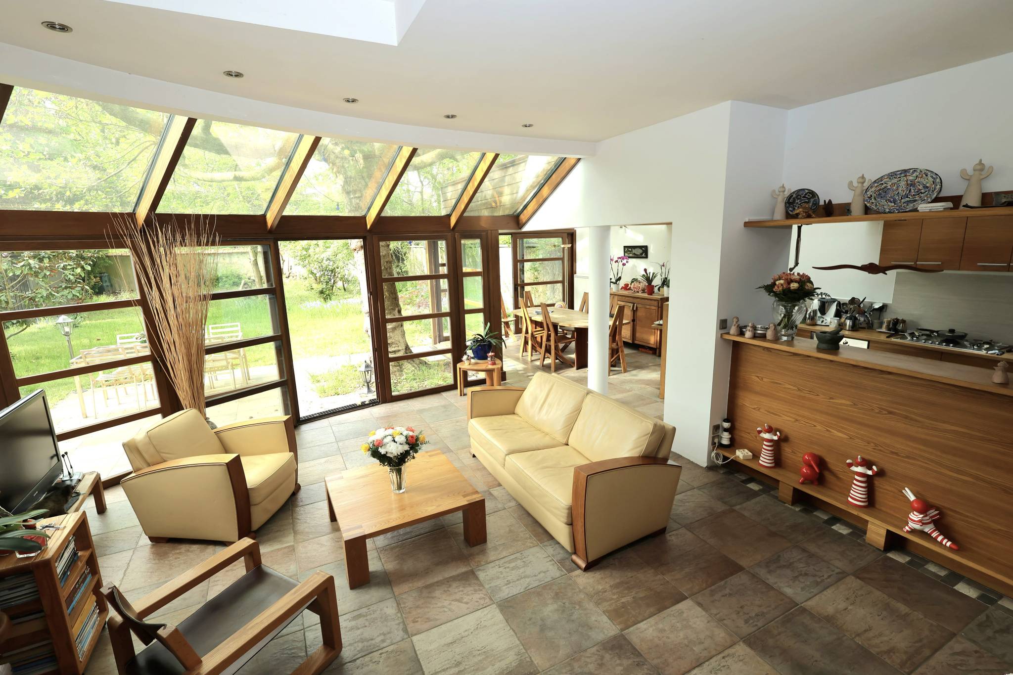 Bright open living room with beige sofas, wooden furniture, and large glass doors opening to a green garden outside.