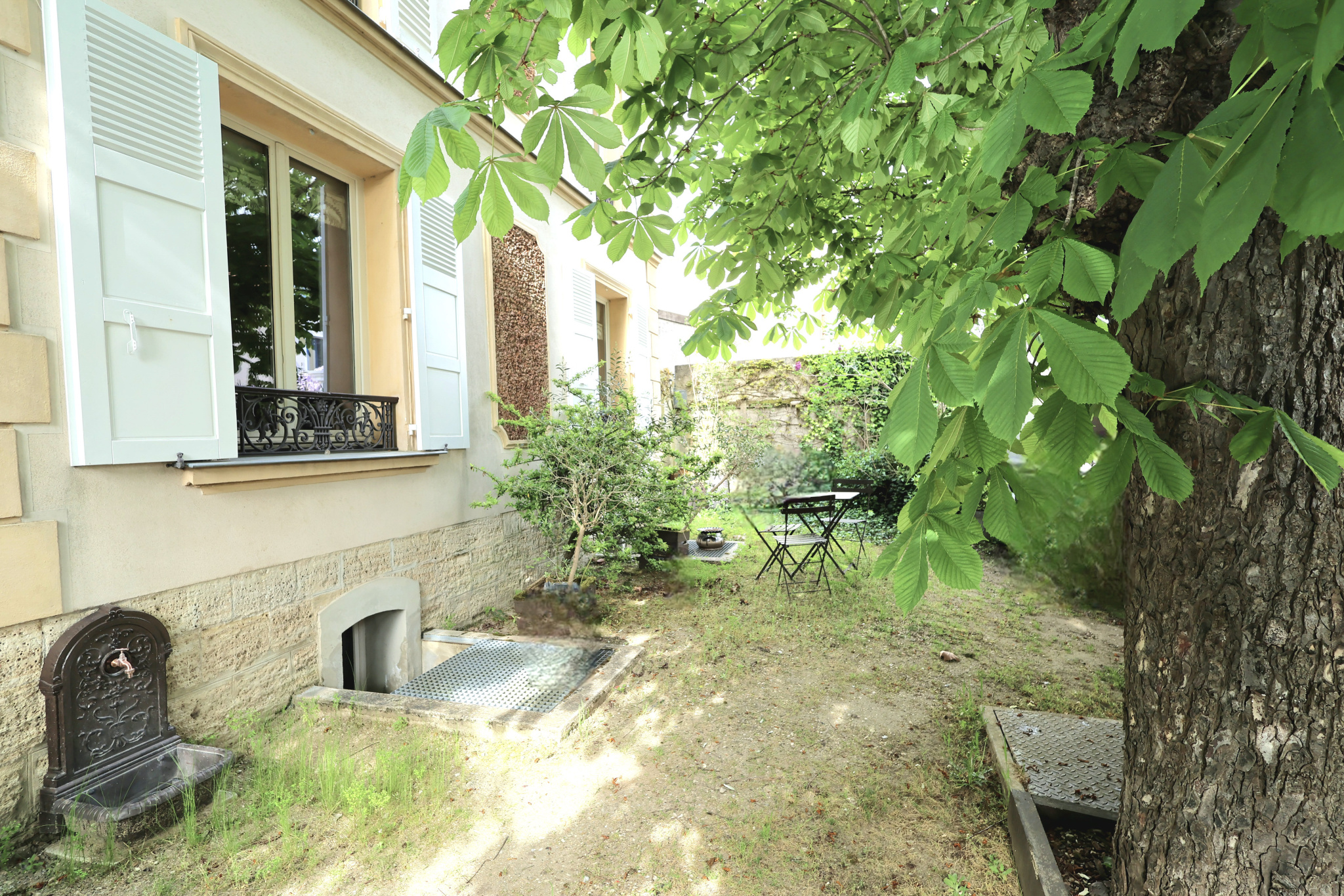 Backyard courtyard beside a beige stone house with pale blue shutters, overlooking a small garden with a black metal bistro set under a large tree.