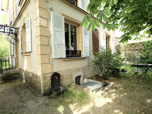 Quaint house exterior with beige walls, light blue shutters, and a stone foundation; small garden with a metal table and chairs under a leafy canopy.