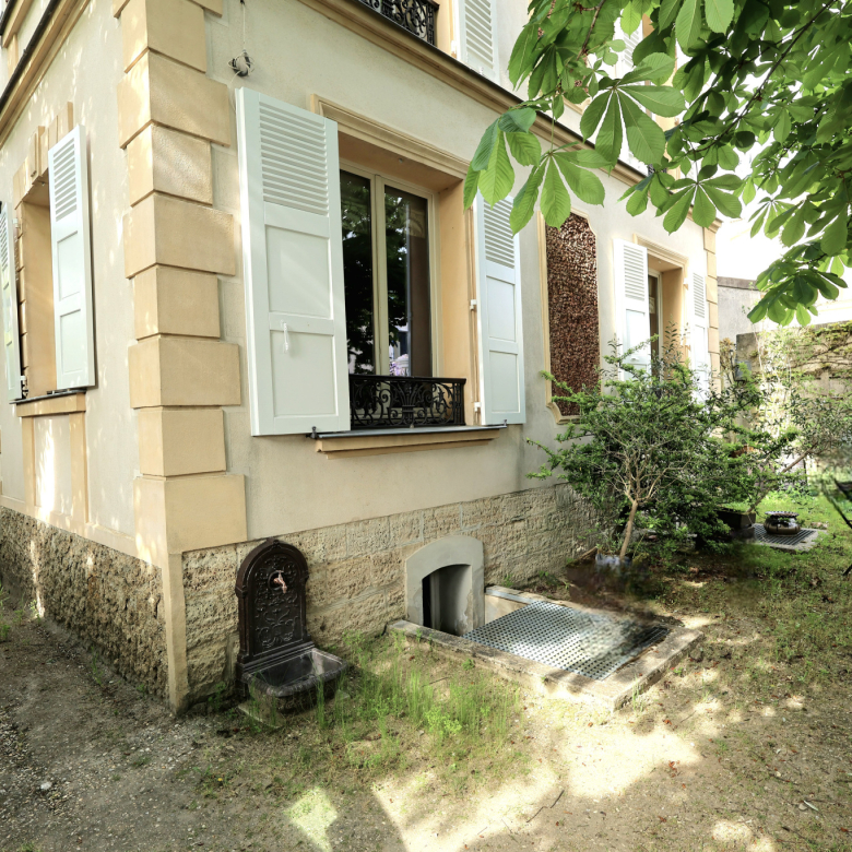Quaint house exterior with beige walls, light blue shutters, and a stone foundation; small garden with a metal table and chairs under a leafy canopy.