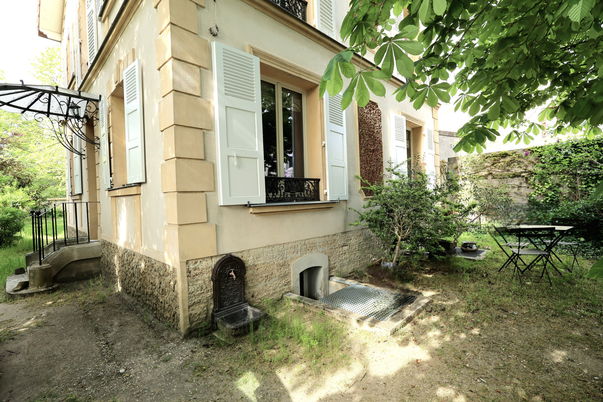 Quaint house exterior with beige walls, light blue shutters, and a stone foundation; small garden with a metal table and chairs under a leafy canopy.