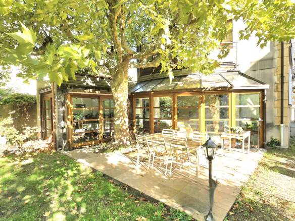 Sunlit patio with white chairs around a long table under a large tree beside a glass-walled conservatory.