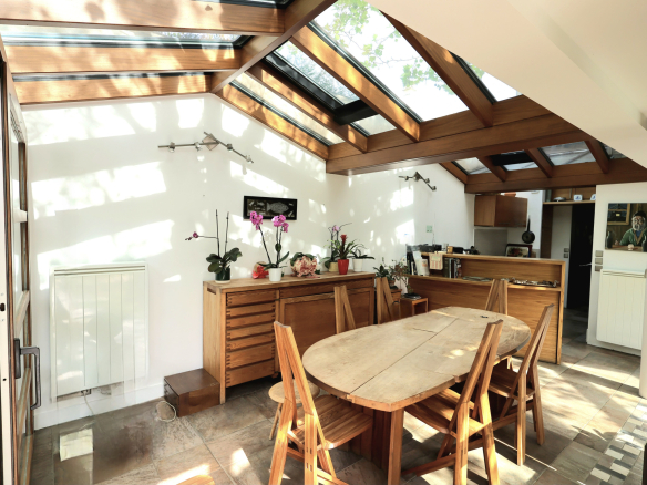 Bright dining room with a round wooden table and six chairs under a skylit, timber-beamed ceiling; potted plants on a sideboard and a kitchen area in the background.