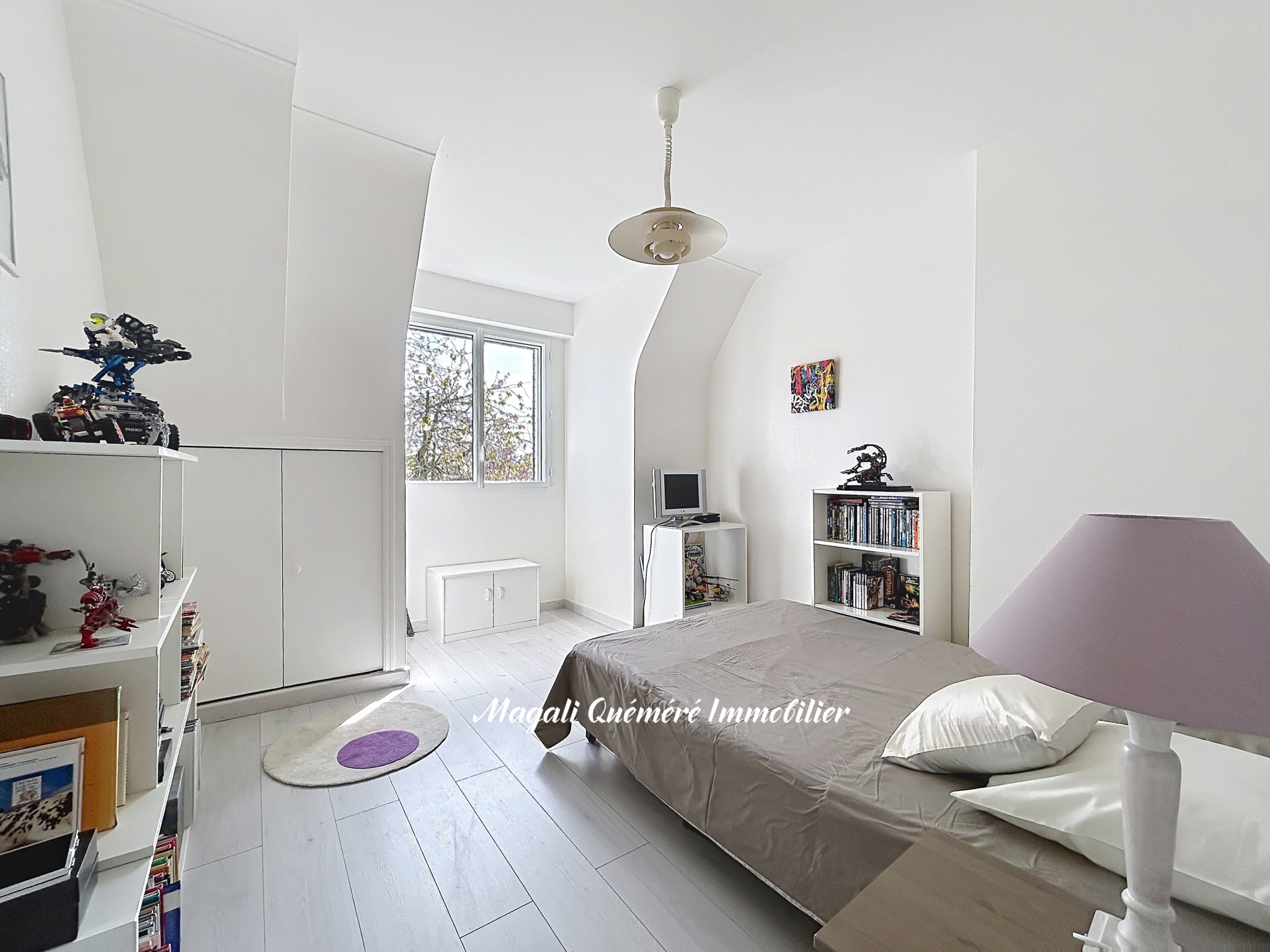 Bright white bedroom with a low bed, purple lamp, and white shelves full of toys and books, near a window showing trees outside.