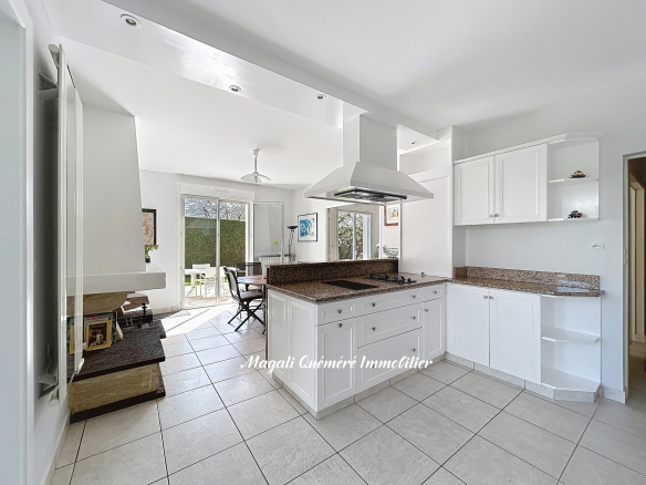 Bright white kitchen with granite countertops and a central island hosting a cooktop and range hood; dining area and sliding doors to the patio visible in the background.