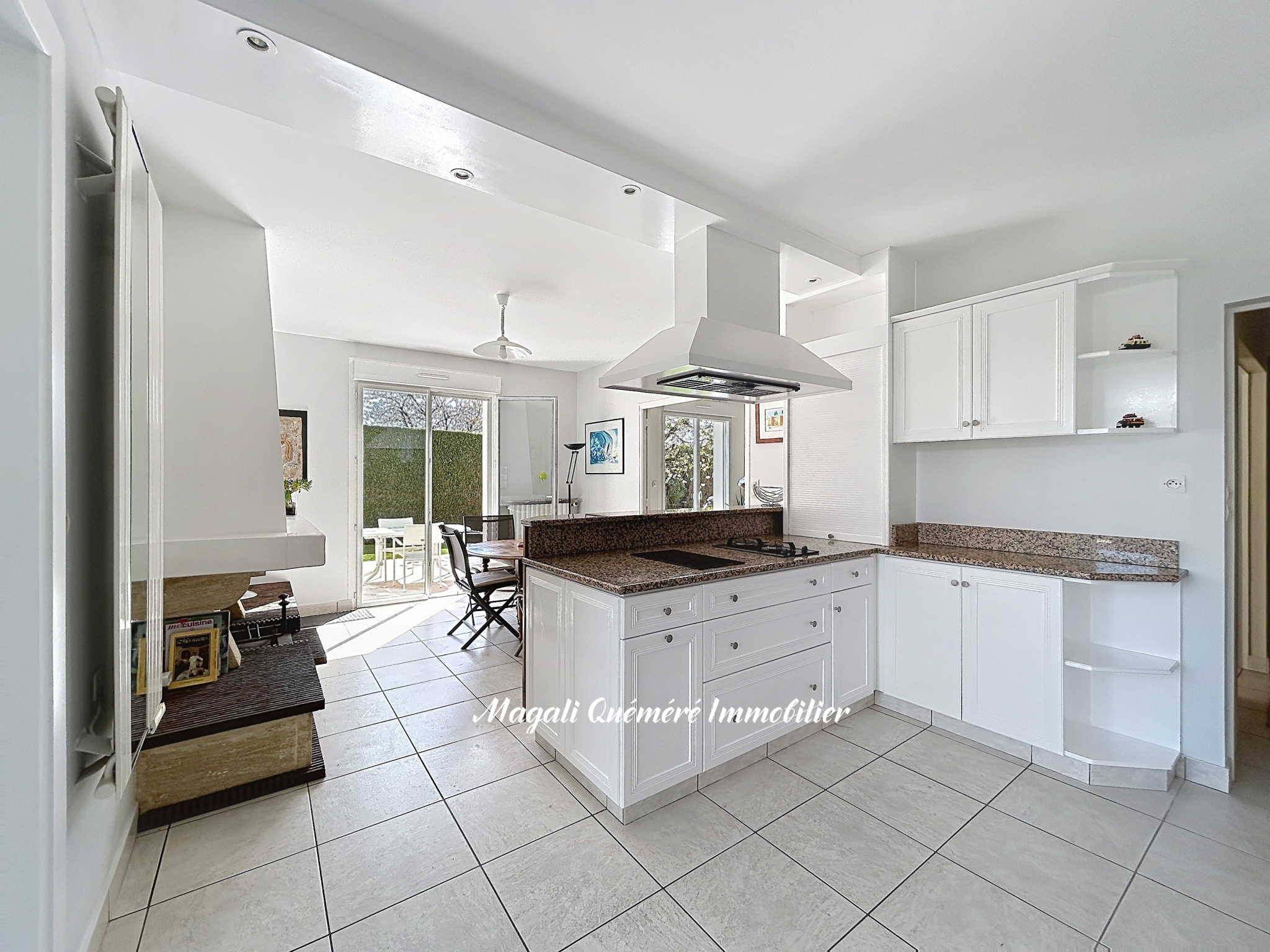 Bright white kitchen with granite countertops and a central island hosting a cooktop and range hood; dining area and sliding doors to the patio visible in the background.