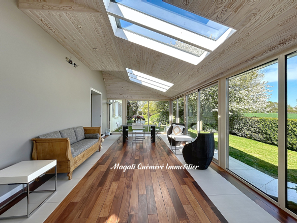 Bright sunroom with a wooden floor, large glass walls and skylights; a grey sofa and wicker chair set facing a green garden.