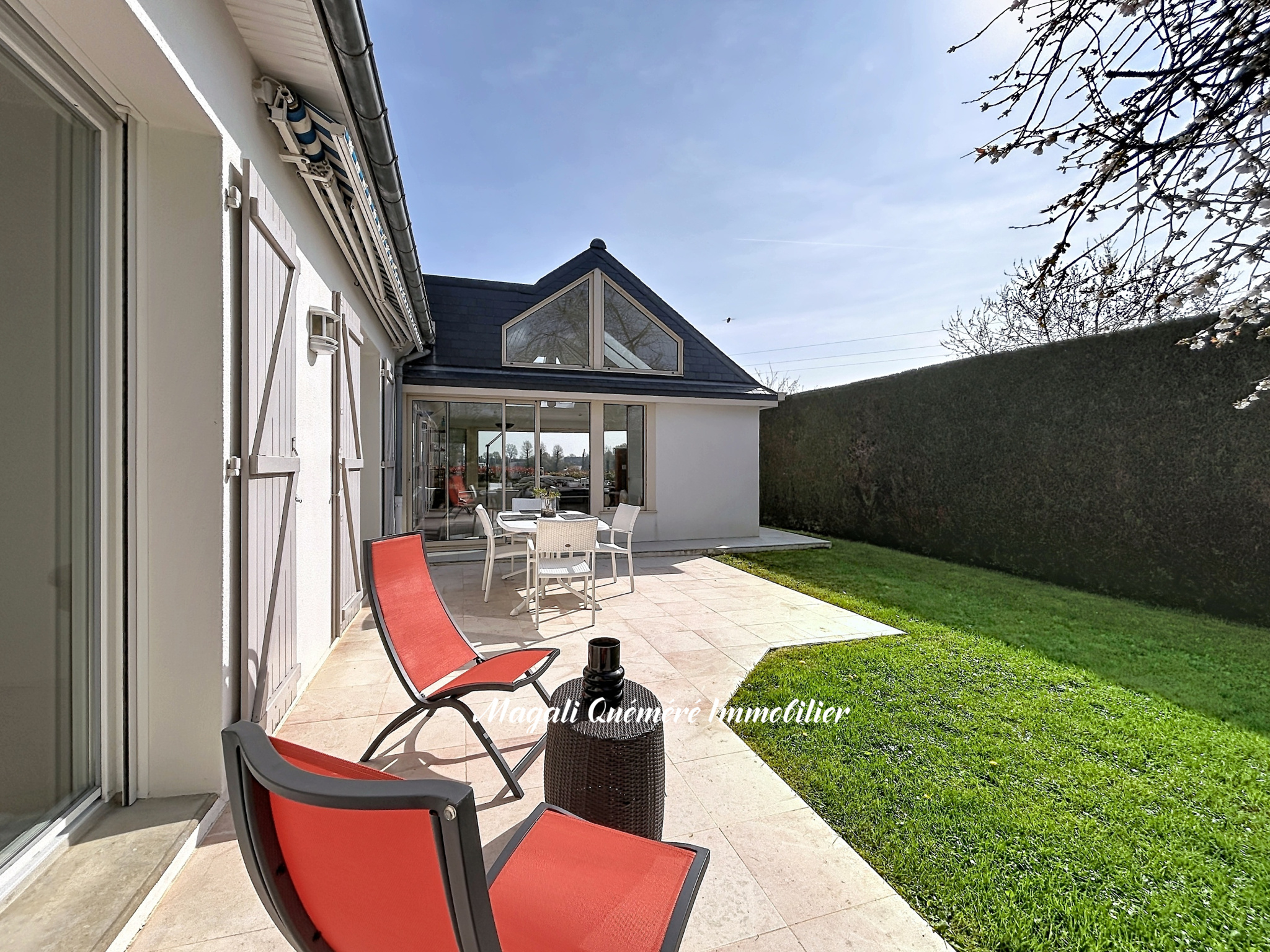 Sunlit backyard patio with red lounge chairs around a small table, white house with glass doors behind them.