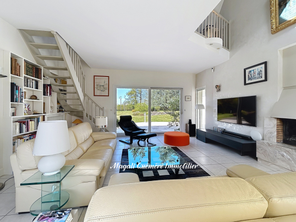 Bright modern living room with white leather sofas, glass coffee table, and built-in bookshelves under a white staircase, opening to a sunny backyard through sliding doors and a large window.