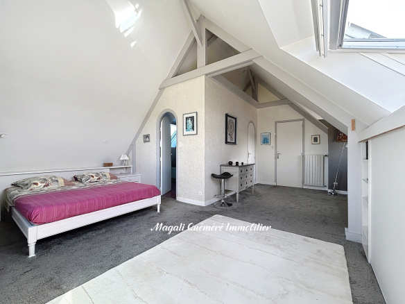 Attic bedroom with white sloped ceilings, a white bed with pink bedding, and a dresser area under a skylight. Watermark reads Magali Quéméré Immobilier in front.