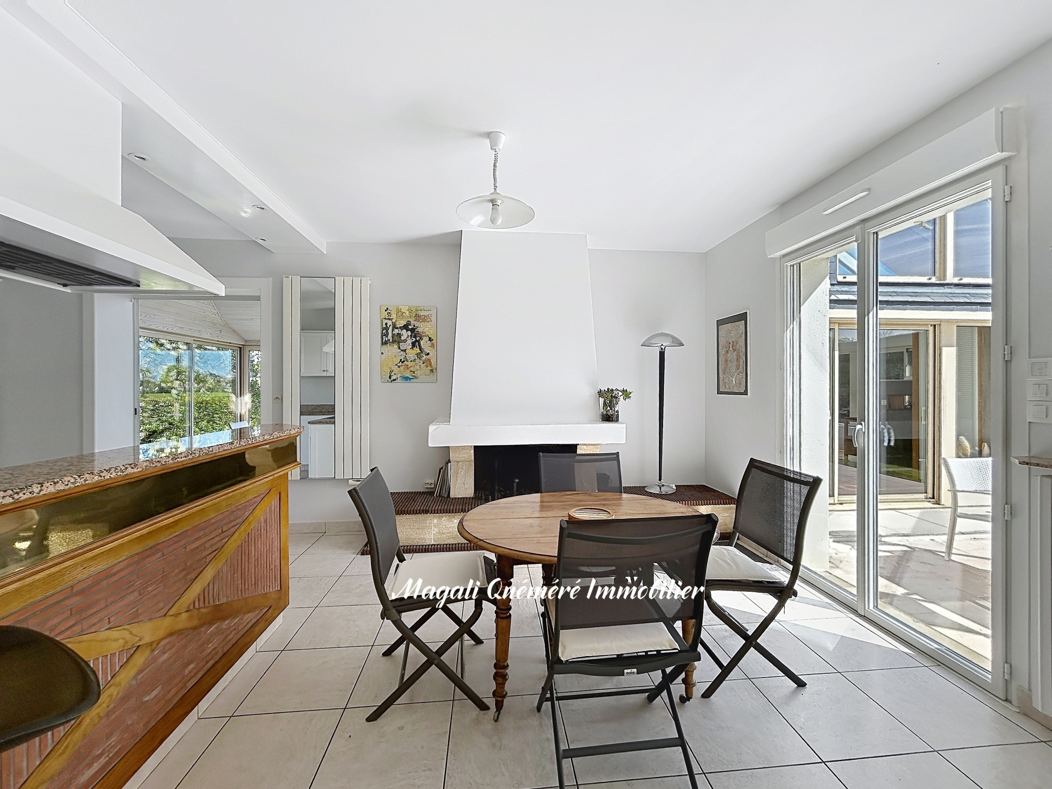 Bright open-plan living and dining area with a white fireplace, round wooden table, and four black chairs; sliding doors to an outdoor patio on the right.
