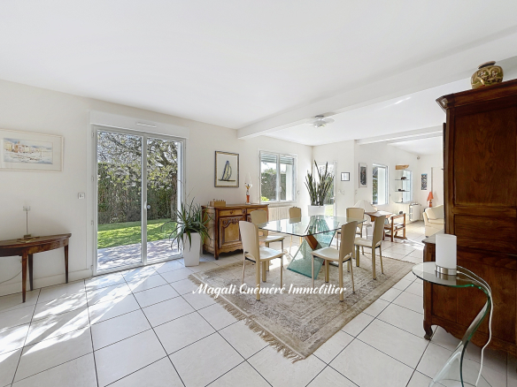 Bright dining room with a glass rectangular table surrounded by cream chairs, rug, and plants; sliding doors open to a garden.