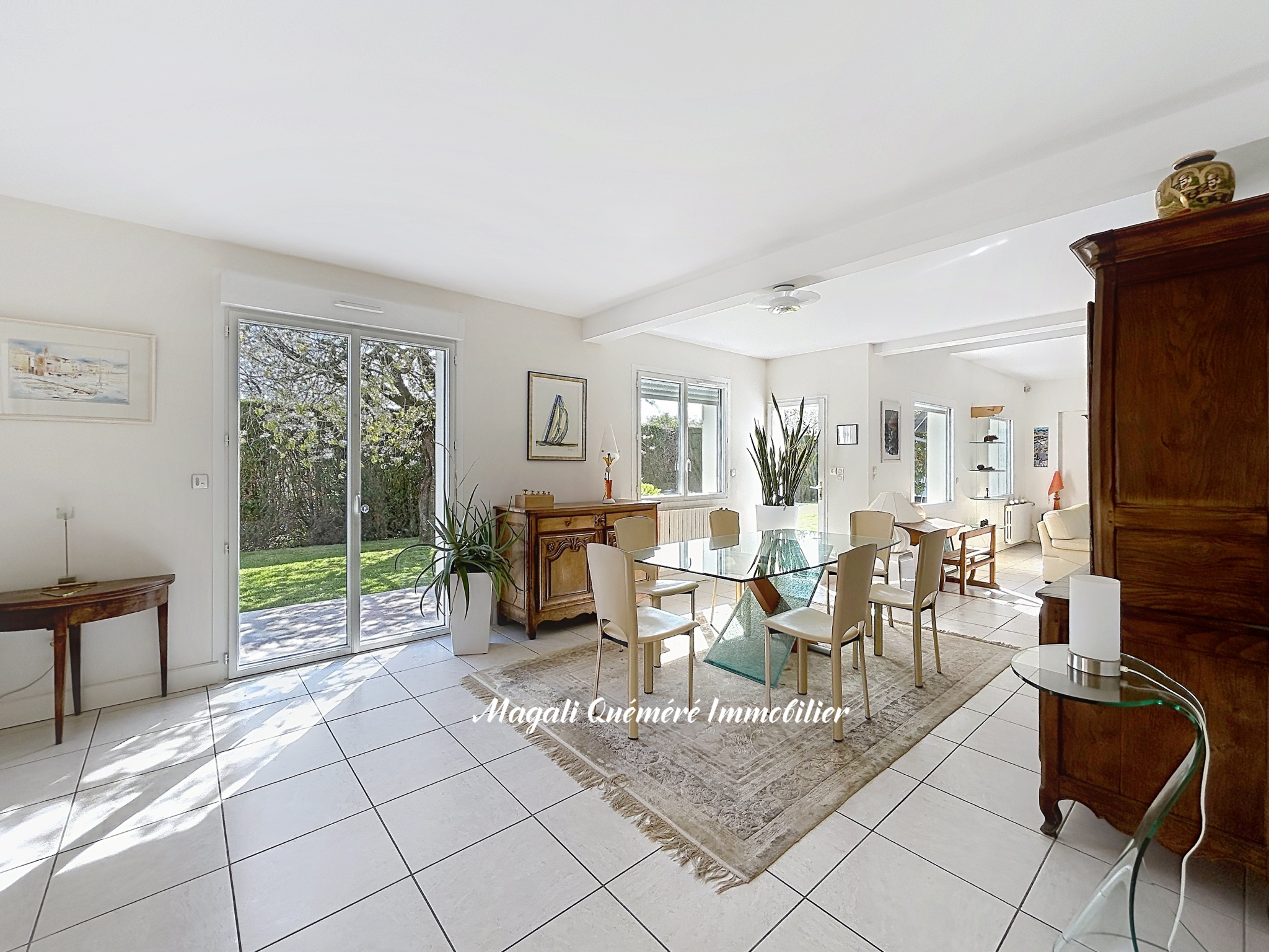 Bright dining room with a glass rectangular table surrounded by cream chairs, rug, and plants; sliding doors open to a garden.