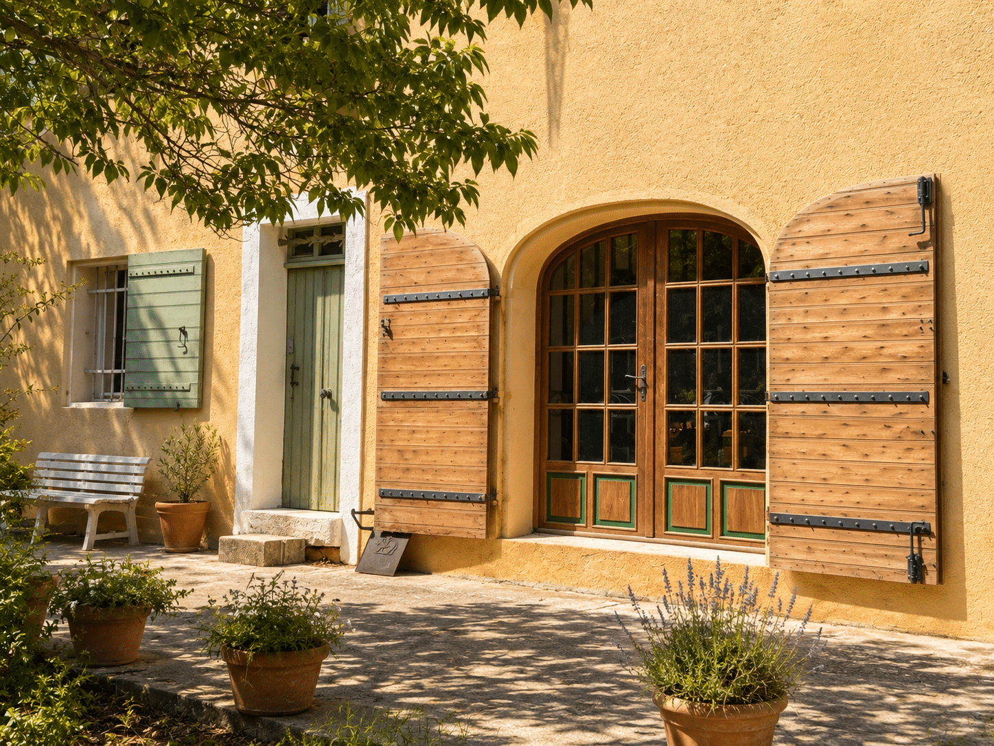 Entrée d'un mas provençal avec volets en bois et portes vitrées, entouré de plantes et d'une allée.