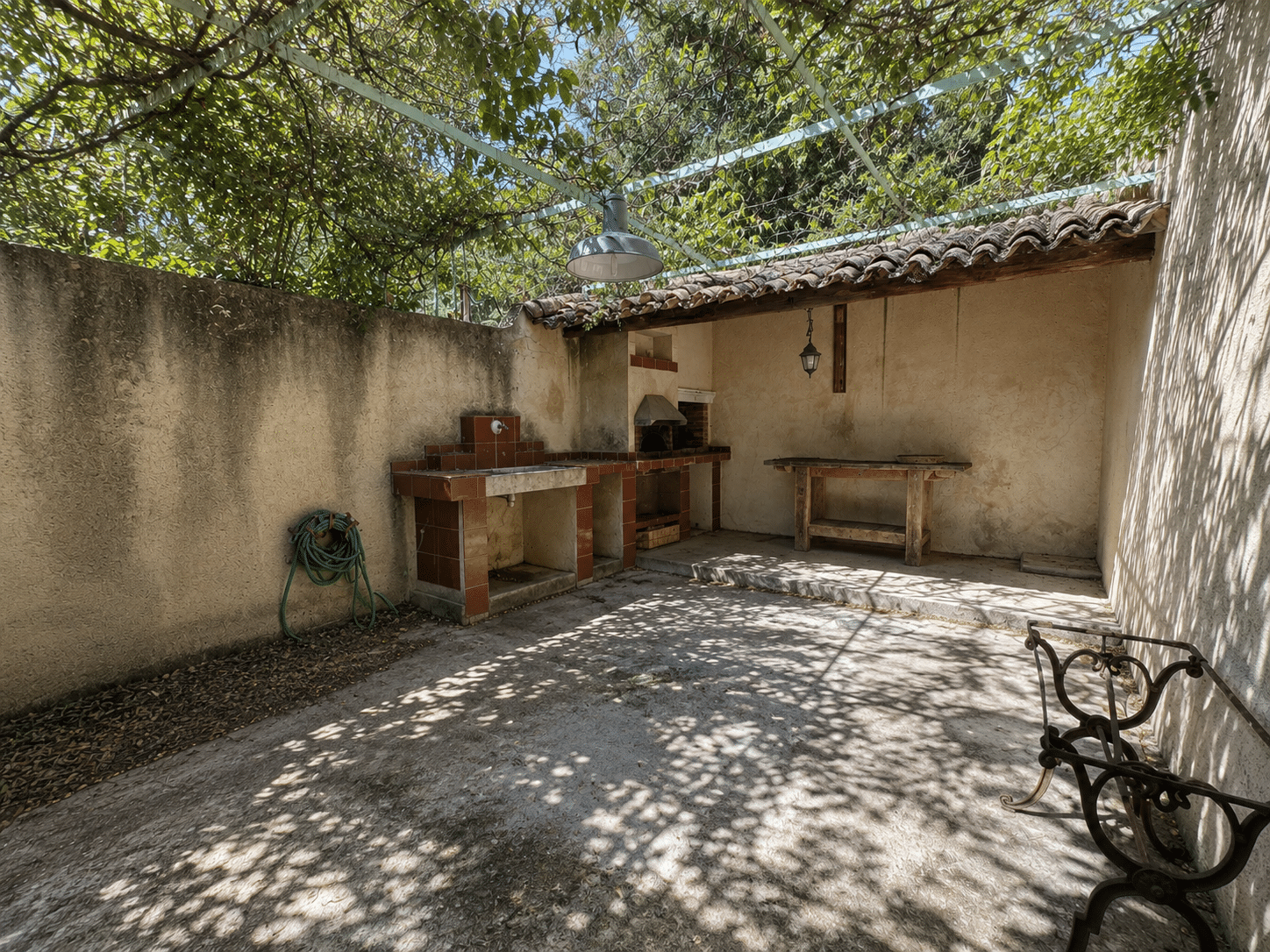 Espace extérieur d'un mas provençal avec barbecue et table en bois, ombragé par des arbres.