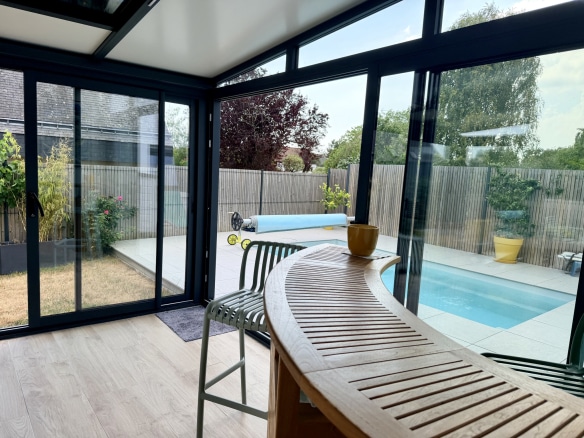 Intérieur d'une véranda lumineuse avec vue sur un jardin et une piscine.