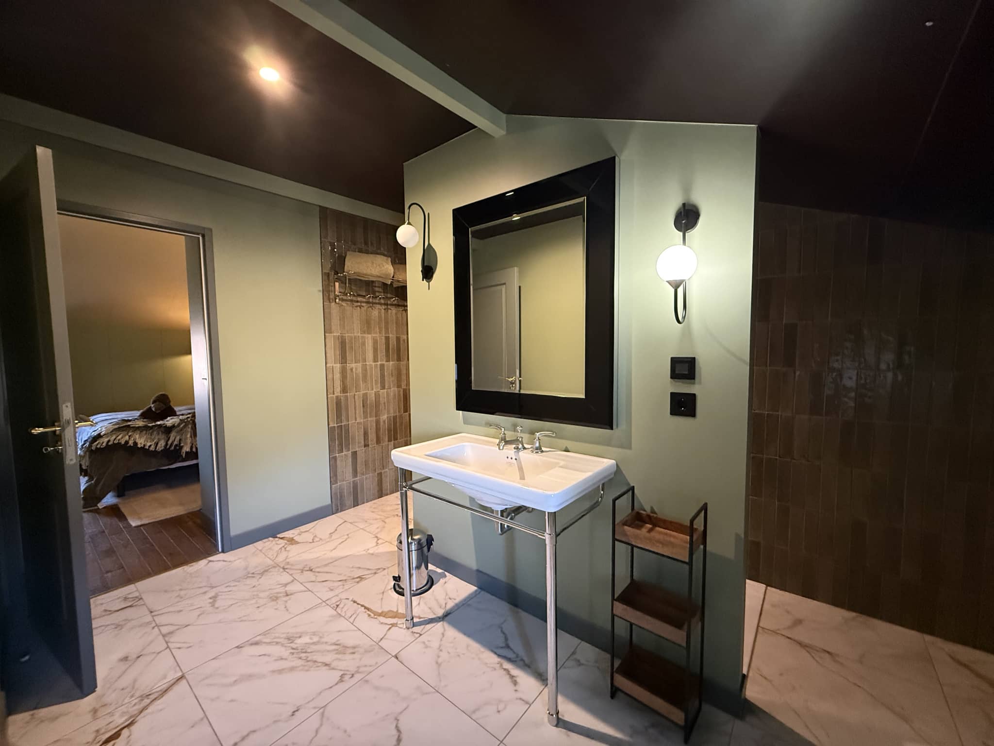Salle de bain moderne avec lavabo sur pied et miroir, chambre à coucher visible en arrière-plan.