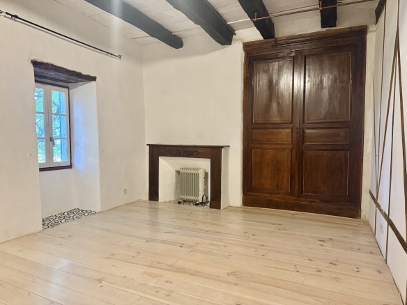 Interior of a stone house with a wooden floor and a large wooden door, windows visible.