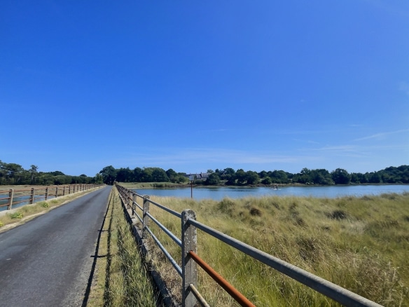 Vue d'un chemin bordé de clôtures menant à une propriété historique près de Pont-l'Abbé, avec des champs et un étang.