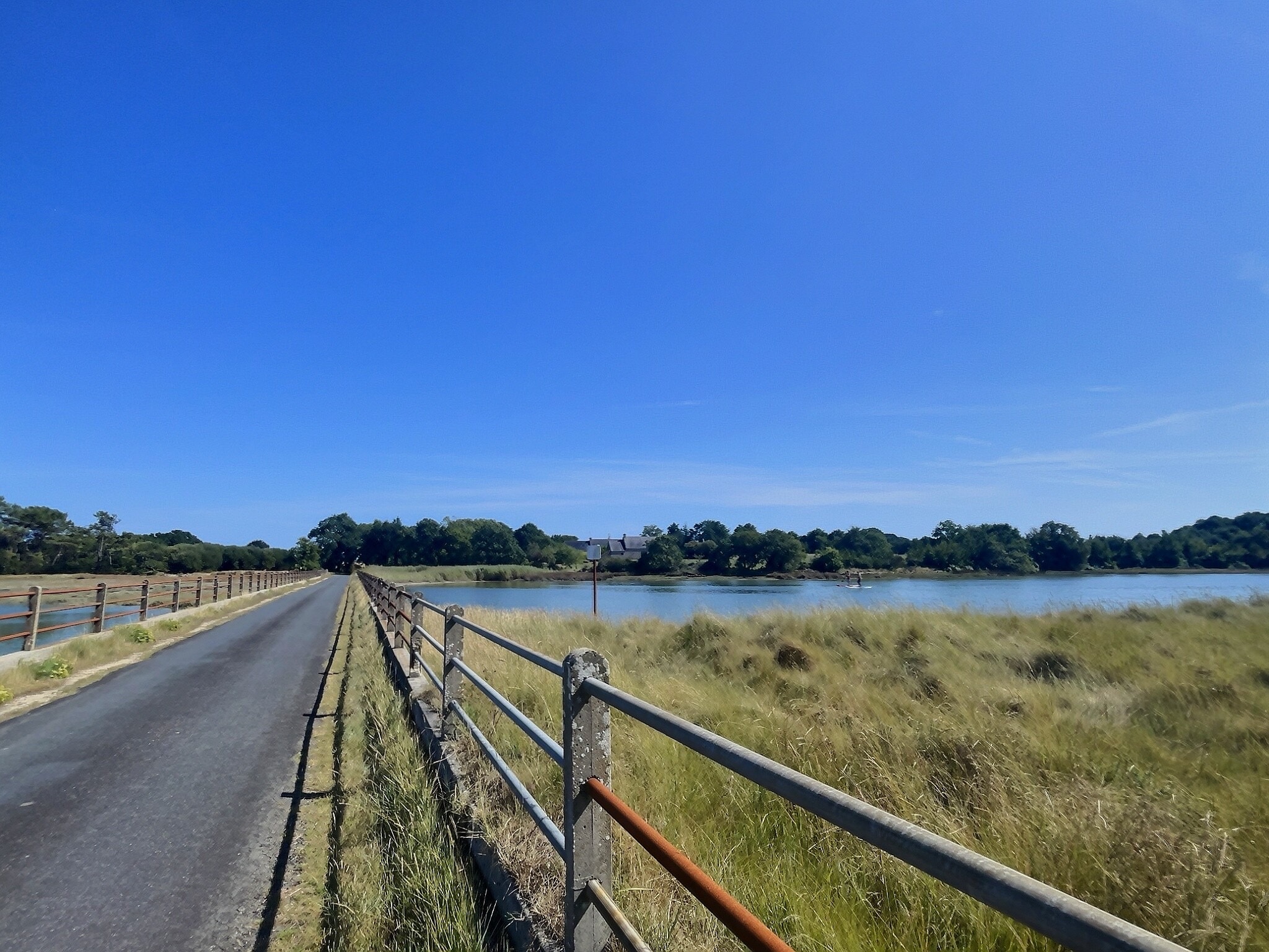 Vue d'un chemin bordé de clôtures menant à une propriété historique près de Pont-l'Abbé, avec des champs et un étang.