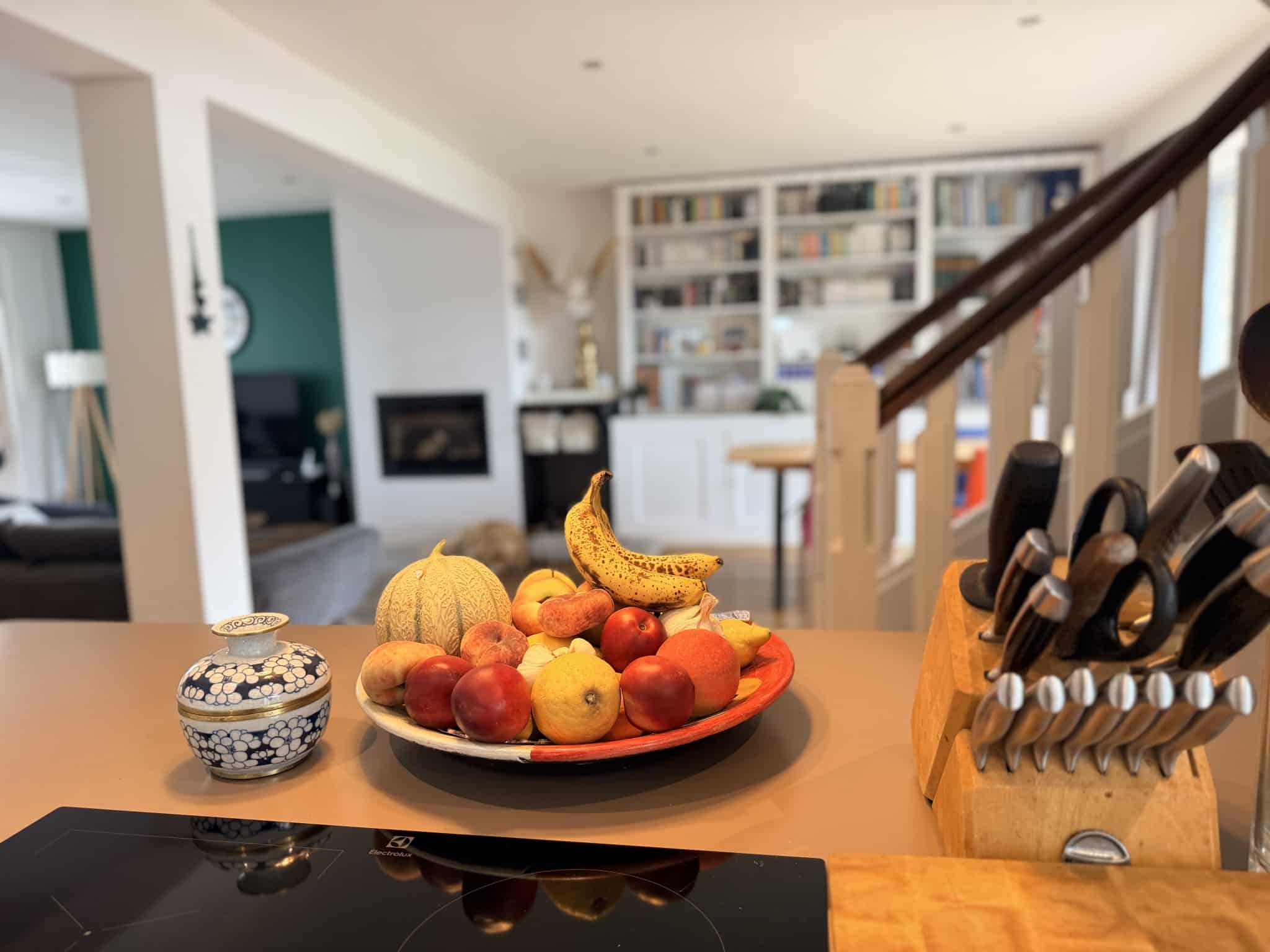 Intérieur d'une maison avec un plateau de fruits sur une table, vue sur un salon et une bibliothèque en arrière-plan.