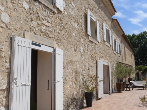 Façade en pierre d'un mas avec portes blanches, terrasse en carrelage et plantes en pot.