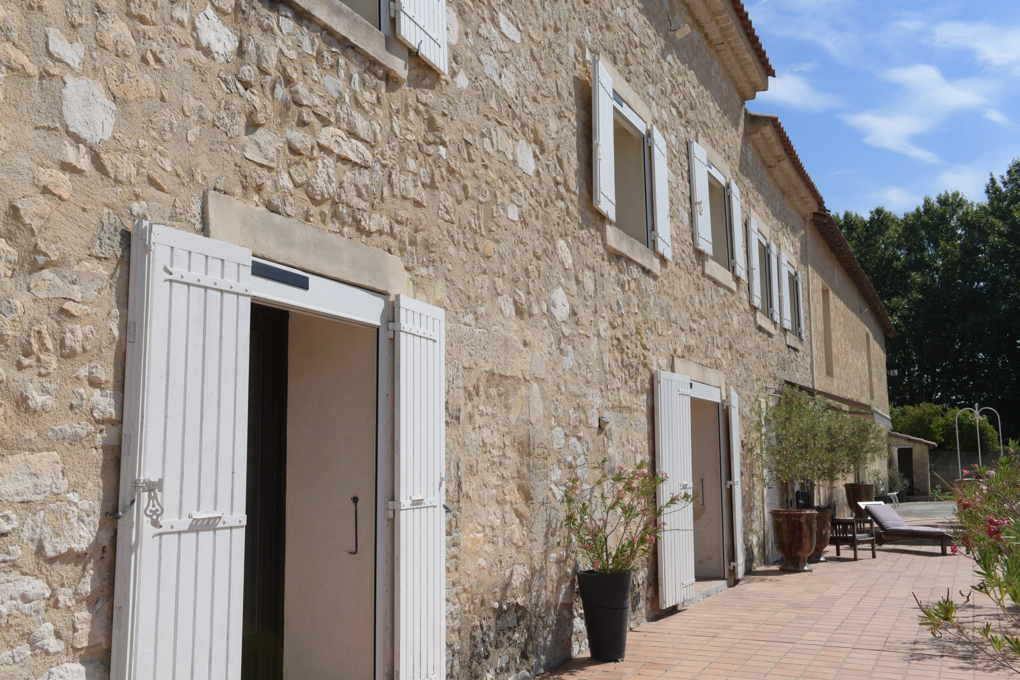 Façade en pierre d'un mas avec portes blanches, terrasse en carrelage et plantes en pot.