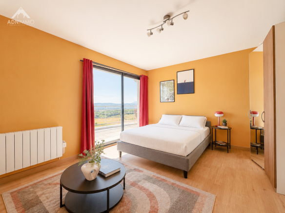 Chambre lumineuse avec lit double et vue sur la nature, murs orange et fenêtres grandes.