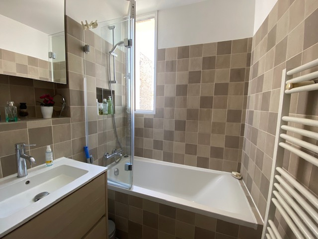 Salle de bain d’un appartement à Paris 12ème Salle de bain moderne avec baignoire et douche, carrelage beige, fenêtre laissant entrer la lumière.