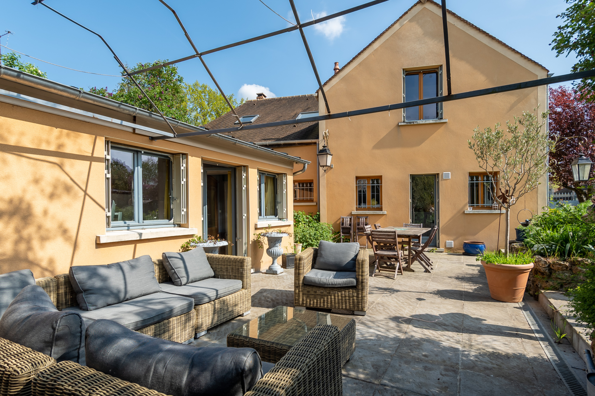 Terrasse d'une maison avec mobilier en osier, jardin verdoyant et façade jaune.
