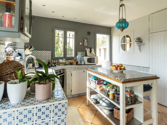 Cuisine lumineuse avec carrelage et îlot central, plantes en pot, et fenêtres donnant sur l'extérieur.