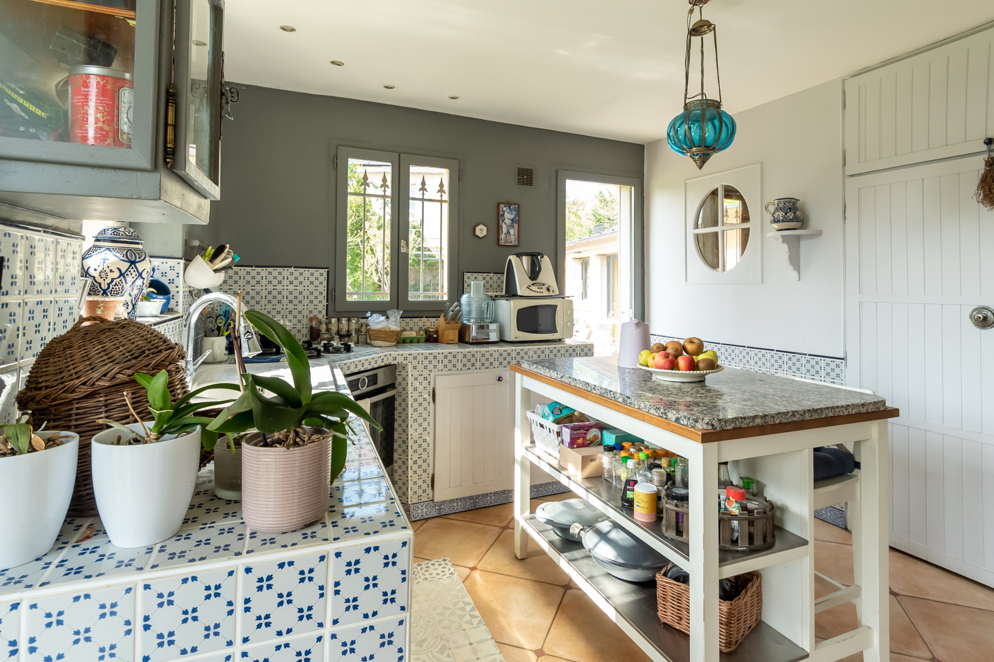 Cuisine lumineuse avec carrelage et îlot central, plantes en pot, et fenêtres donnant sur l'extérieur.