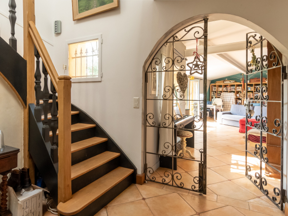Intérieur d'une demeure de charme avec un escalier en bois et une porte en fer forgé menant à un séjour lumineux.