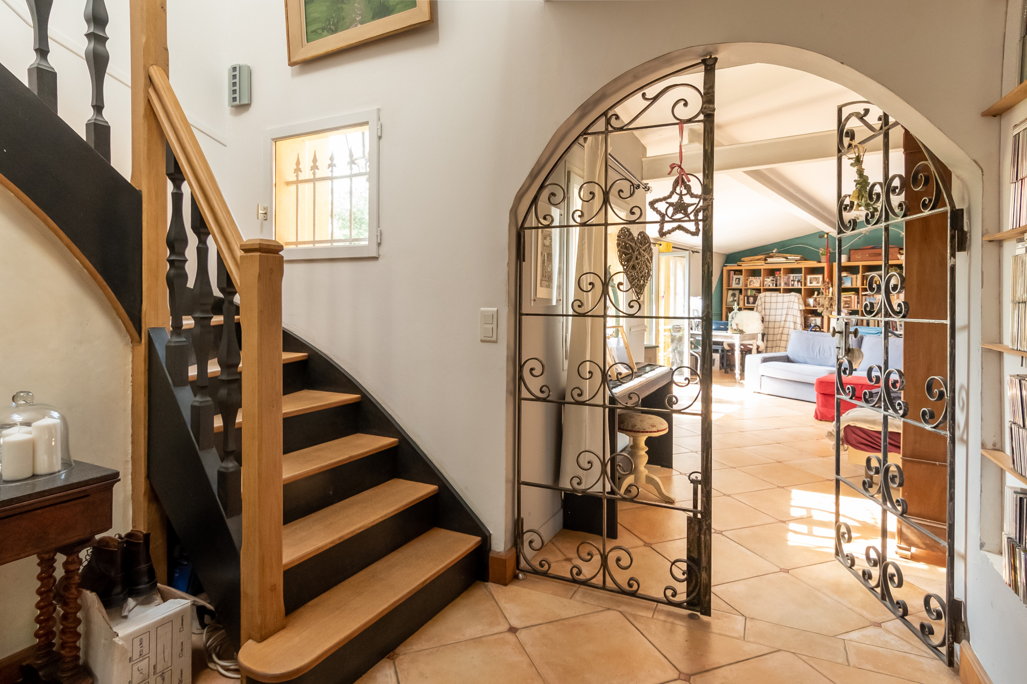 Intérieur d'une demeure de charme avec un escalier en bois et une porte en fer forgé menant à un séjour lumineux.