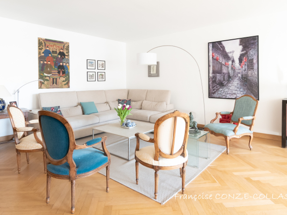 Salon lumineux d'un appartement familial avec canapé, table en verre et chaises anciennes.