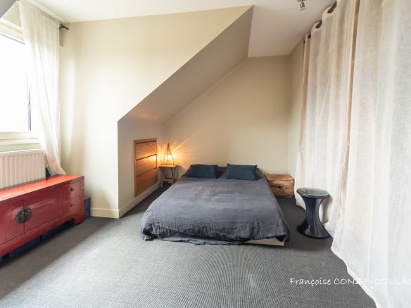 Chambre avec lit au sol, décorée sobrement, murs inclinés et fenêtre laissant entrer la lumière.
