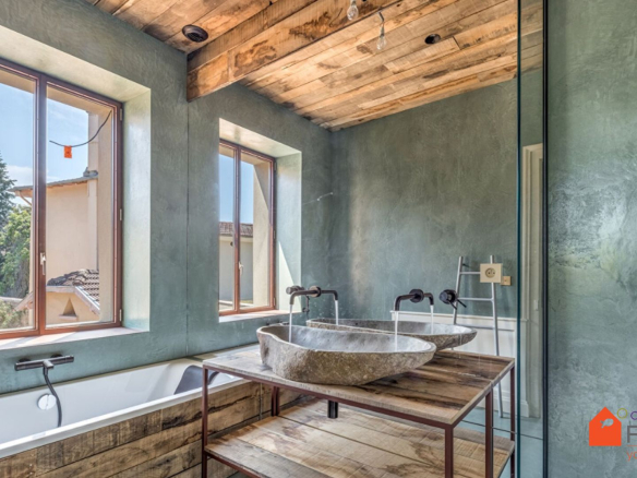 Salle de bain moderne avec baignoire et vasques en pierre, murs en béton ciré et fenêtres lumineuses.