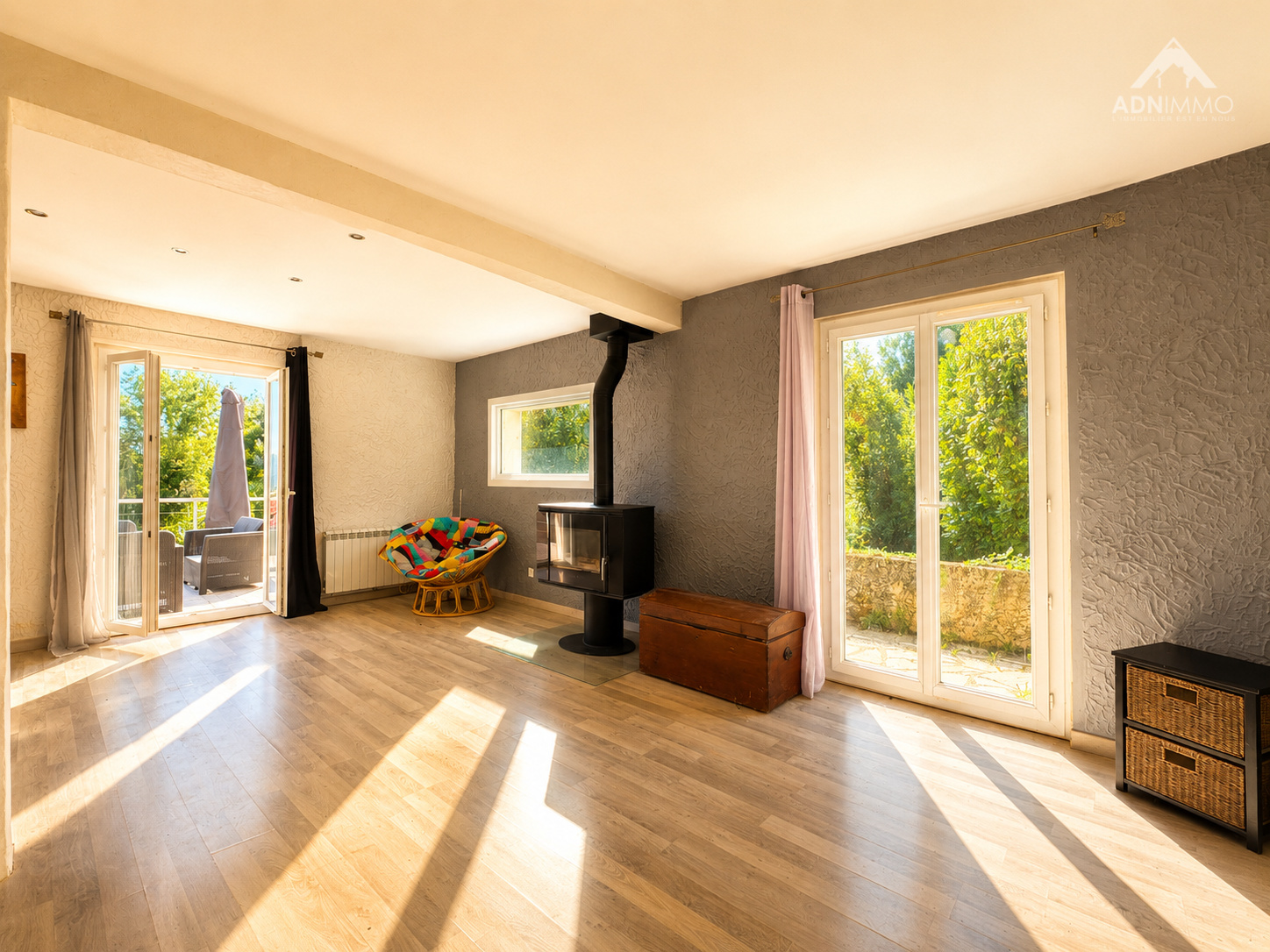 Intérieur lumineux d'une maison avec poêle à bois et grandes fenêtres donnant sur un jardin.
