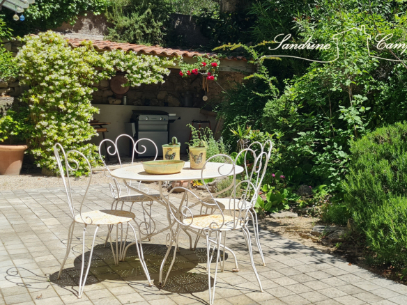 Terrace with table and chairs in a green garden, surrounded by plants and trees.