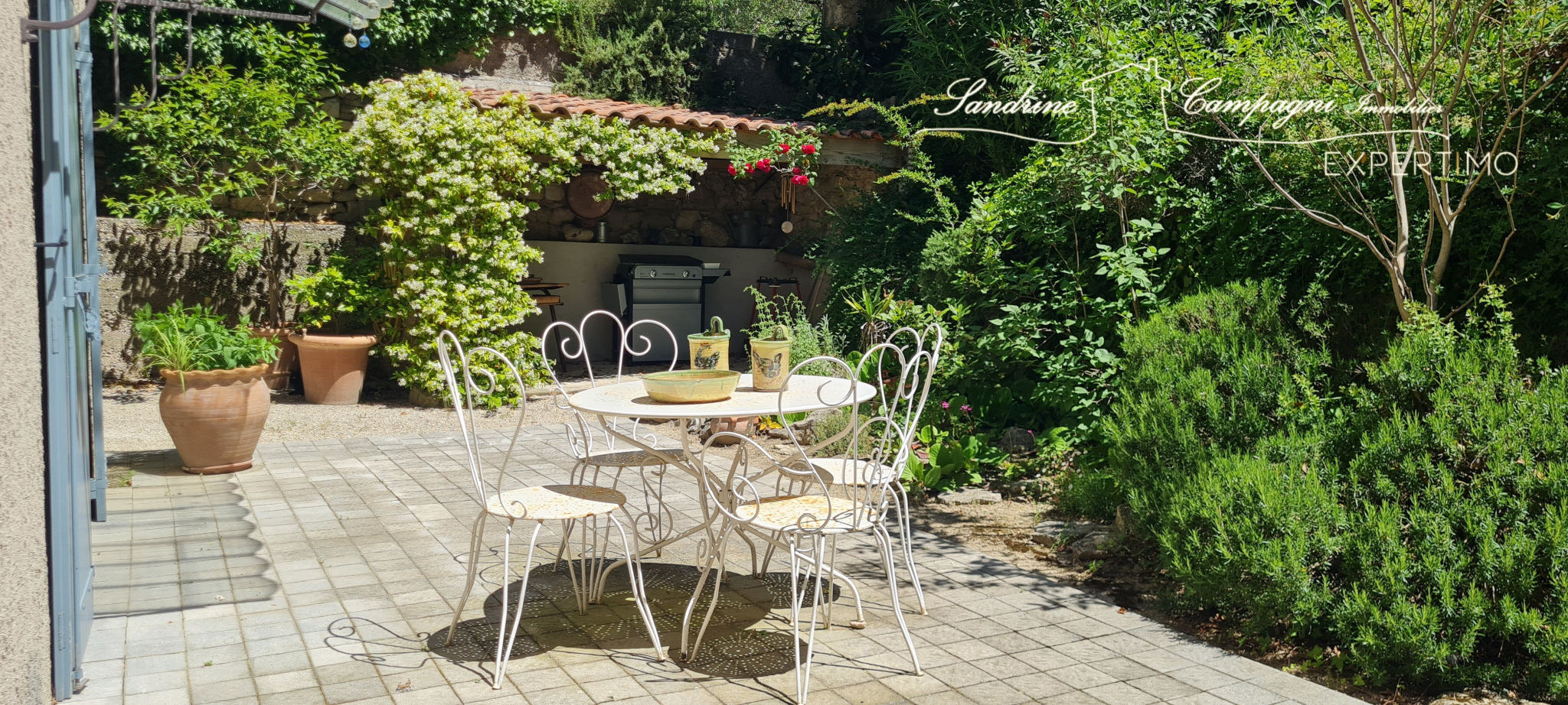 Terrace with table and chairs in a green garden, surrounded by plants and trees.